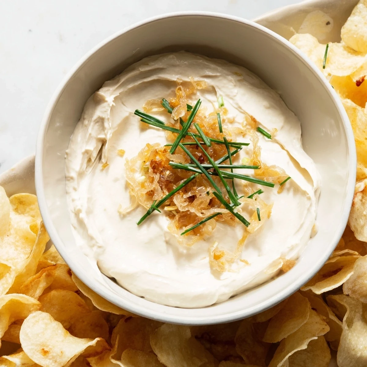 A close-up of a bowl of homemade onion dip and a pile of salty potato chips for dipping.